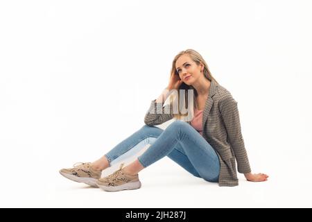 Femme blonde jeune adulte en Jean et blazer à carreaux assis au sol sur fond blanc et regardant avec un sourire doux. Prise de vue en studio avec espace de copie isolé. Photo de haute qualité Banque D'Images