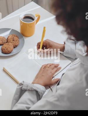 Une femme écrit ses pensées dans un journal du matin dans la cuisine. Banque D'Images