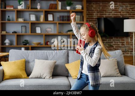 Bonne petite fille portant un casque rouge dansant et chantant à l'aide d'un smartphone comme un microphone. Enfant mignon Profitez de votre musique préférée Banque D'Images