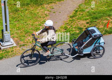 Une jeune mère fait du vélo avec son enfant dans un transporteur le long d'une piste cyclable à Steveston, en Colombie-Britannique, au Canada Banque D'Images