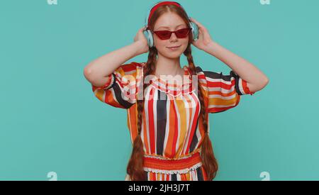 Portrait d'une fille à tête rouge en robe écouter de la musique via des écouteurs et danser disco berner autour d'avoir plaisir expressif gesticaling mains. Jeune adolescent sur fond de mur bleu de studio Banque D'Images