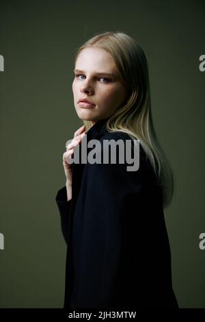 Un portrait rapproché d'une blonde regardant par-dessus son épaule dans une veste noire posant dans un studio sur un fond vert. Le concept de mode élégant Banque D'Images