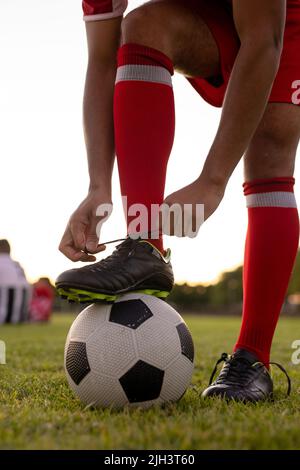 Coupe basse d'athlète de sexe masculin caucasien portant des chaussettes rouges avec une jambe sur le ballon de football nouant le lacet Banque D'Images