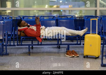 Homme africain attendant le vol avec un smartphone. Un homme noir allongé sur un banc près des bagages à l'aéroport Banque D'Images