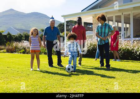 Famille multiraciale de plusieurs générations regardant un garçon jouant au football sur un terrain herbeux dans la cour en été Banque D'Images