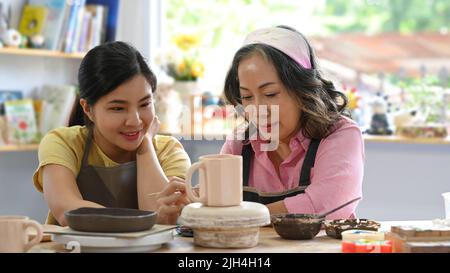 Jolie femme asiatique et femme mûre appréciant le processus créatif de la coloration de poterie dans l'atelier de poterie Banque D'Images