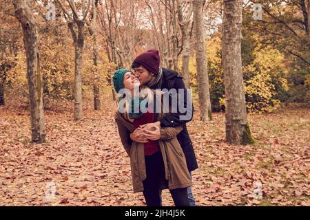 Blanc homme caucasien embrassant sa petite amie de derrière lui donnant un baiser sur la joue tandis qu'elle est souriante dans un parc en automne avec le sol plein de Banque D'Images