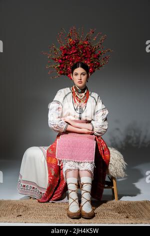 pleine longueur de jeune femme ukrainienne en vêtements traditionnels et couronne florale rouge assise sur un banc gris Banque D'Images