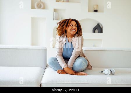 Jeune fille millénaire afro-américaine, heureuse et heureuse, asseyez-vous sur un canapé confortable dans un salon moderne et confortable. Profitez de sa vie Banque D'Images