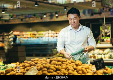 un jeune homme asiatique choisit et cueille dans un sac écologique légumes pomme de terre ou fruits dans le supermarché. un client de sexe masculin se tient dans une épicerie près du comptoir b Banque D'Images