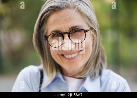 Gros plan portrait de charmante femme mûre de cheveux gris en lunettes debout à l'extérieur appréciant des vacances à temps libre voyager autour du monde à la retraite. Femme mûre avec une peau parfaite portant une chemise bleue. Banque D'Images