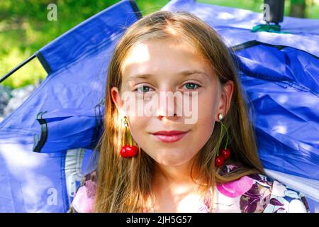 Refocalisation belle blonde jeune fille avec cerise douce sur l'oreille. Belle adolescente souriante à proximité, contre le vert du parc d'été. Tente de camping. Enfant Banque D'Images