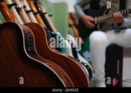 Beaucoup de guitares électriques et acoustiques sur un stand au premier plan avec un musicien jouant en arrière-plan. Corps des guitares. Guitares sur Music Store. Musicien jouant un fils en arrière-plan avec une guitare noire. Banque D'Images