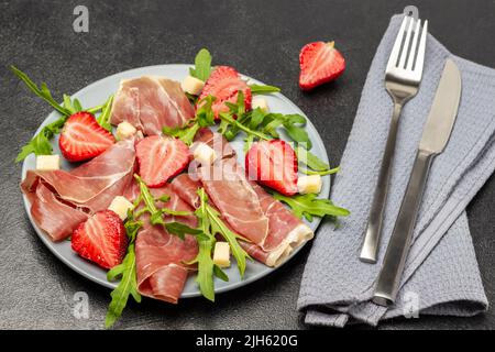 Fourchette et couteau sur serviette grise. Jamon espagnol avec fromage, fraises et arugula sur plaque grise. Vue de dessus. Arrière-plan noir Banque D'Images