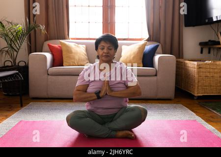 Pleine longueur de biracial mature femme méditant en position de prière sur le tapis dans la salle de séjour à la maison Banque D'Images