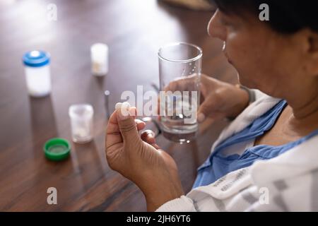 Vue en grand angle de la femme biraciale mature tenant des médicaments et de l'eau potable à la table à la maison Banque D'Images