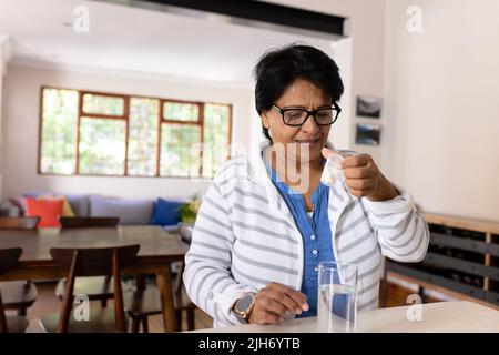 Biracial mature femme avec de l'eau potable sur la table portant des lunettes de vue vérifiant la bouteille de médicament Banque D'Images