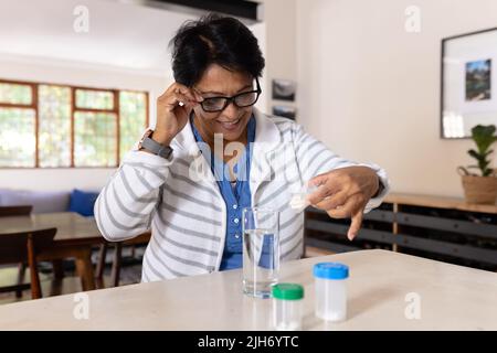 Souriante biracial femme mature portant des lunettes de vue vérifiant les bouteilles de médicaments sur la table à la maison Banque D'Images
