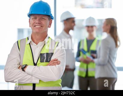 IBE a construit une grande équipe au fil des ans. Portrait court d'un beau ouvrier de construction mature debout avec ses bras pliés et ses collègues Banque D'Images
