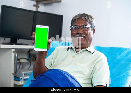 Heureux patient souriant montrant le téléphone mobile à écran vert en regardant la caméra pendant le sommeil sur le lit à l'hôpital - concept de service de santé Banque D'Images