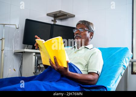 Homme âgé avec livre de lecture de lunettes à l'hôpital pendant le lit - concept de relaxation, de réadaptation et de traitement d'assurance-maladie. Banque D'Images
