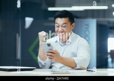 Réussite gagner. Un jeune homme d'affaires asiatique tient le téléphone en main, se réjouit de gagner, bonne nouvelle, lit le message. Sourire. Il est assis dans un bureau moderne à une table avec un ordinateur portable et une tablette Banque D'Images