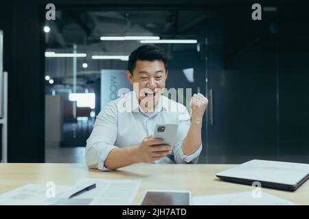 Réussite gagner. Un jeune homme d'affaires asiatique tient le téléphone en main, se réjouit de gagner, bonne nouvelle, lit le message. Sourire. Il est assis dans un bureau moderne à une table avec un ordinateur portable et une tablette Banque D'Images