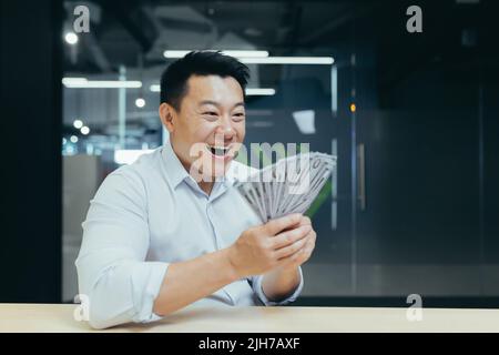 Un jeune homme asiatique heureux a reçu un salaire en espèces. Il tient de l'argent entre ses mains, le montre, se réjouit. Bureau dans un bureau moderne Banque D'Images