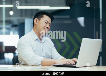 Un jeune homme, un homme d'affaires asiatique au travail au bureau, s'assoit à la table, tient le cou, ressent de la douleur, ferme les yeux, Banque D'Images