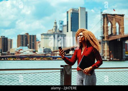 Portrait jeune femme millénaire d'entrepreneur adulte réfléchie avec des lunettes et des cheveux afro dans une conférence téléphonique vidéo à l'extérieur avec Manhattan New York City Skyline derrière le fleuve Hudson Banque D'Images
