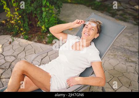 Bonne femme senior mature dans des verres avec le sourire parfait portant totalement blanc relaxant dans jardin d'été en plein air . Concept de mode de vie actif des personnes âgées à la retraite Banque D'Images