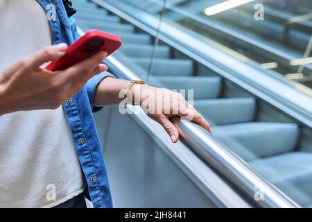 Gros plan d'une main de femme avec un smartphone sur un escalier mécanique, dans un bâtiment moderne Banque D'Images