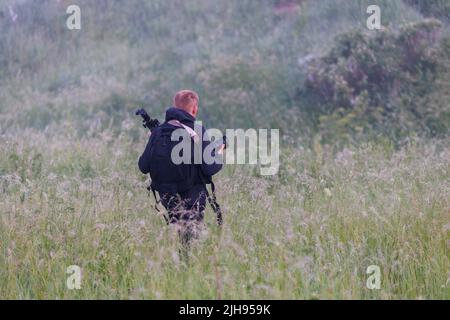 photographe professionnel se déplaçant à travers l'herbe sèche haute dans le matin brumeux à la zone de prise de vue avec l'appareil photo, le trépied et le sac à dos noir Banque D'Images