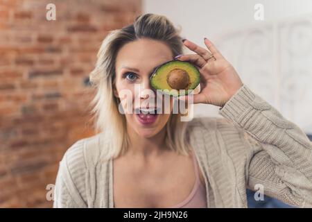 Femme blanche blonde excitée avec avocat délicieux mûr à la maison, moyenne gros plan sain concept. Photo de haute qualité Banque D'Images