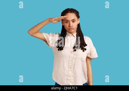 Oui, monsieur. Portrait d'une femme sérieuse responsable avec des dreadlocks noirs saluant commandant, ordre d'écoute avec expression obéissante, portant chemise blanche. Studio d'intérieur isolé sur fond bleu. Banque D'Images