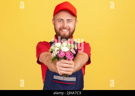 Portrait de l'homme de livraison positif satisfait barbu donnant le bouquet de fleurs, apportant l'ordre, regardant la caméra avec l'expression heureuse. Studio d'intérieur isolé sur fond jaune. Banque D'Images