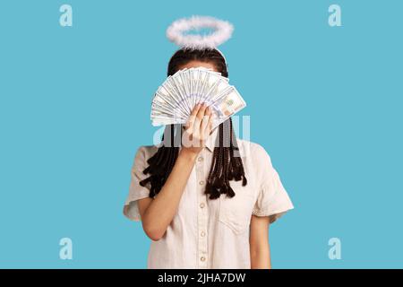 Femme riche et angélique avec des dreadlocks noirs et nimbus à la tête se cachant derrière des billets de banque en dollars, gros profit gagnant de l'argent comptant, portant une chemise blanche. Studio d'intérieur isolé sur fond bleu. Banque D'Images