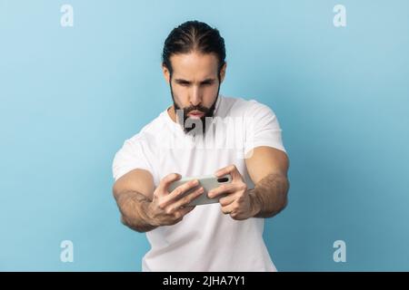 Portrait d'un homme barbu portant un T-shirt blanc debout, utilisant un smartphone et jouant au jeu mobile avec le visage concentré excité. Studio d'intérieur isolé sur fond bleu. Banque D'Images