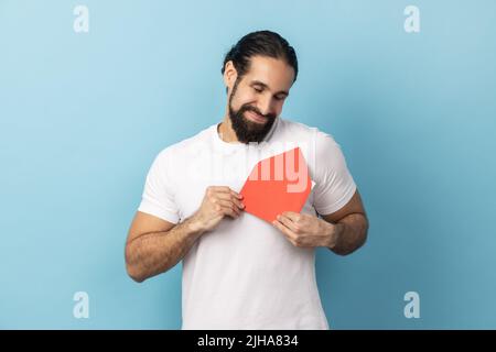 Portrait d'un homme souriant portant un T-shirt blanc debout, tenant une enveloppe rouge enveloppant et gardant les yeux fermés, en appréciant et en étant touché. Studio d'intérieur isolé sur fond bleu. Banque D'Images