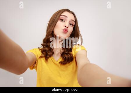 Portrait d'une jeune fille adorable en jaune style décontracté t-shirt prenant selfie, regardant l'appareil photo avec des lèvres pout POV, envoyant l'air baiser. Prise de vue en studio isolée sur fond gris. Banque D'Images