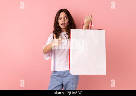 Portrait d'une petite fille choquée portant un T-shirt blanc pointant vers des sacs de shopping, regardant l'appareil photo avec une expression faciale étonnante. Studio d'intérieur isolé sur fond rose. Banque D'Images
