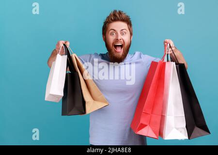 Portrait d'un bel homme barbu excité et stupéfié tenant des sacs de papier de shopping, heureux et heureux avec des réductions de centre commercial, de bons achats. Studio d'intérieur isolé sur fond bleu. Banque D'Images