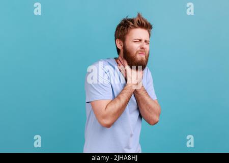 Portrait d'un homme barbu grimaquant touchant le cou, se sentant mal en avalant, mal de gorge, risque d'étouffement ou choc anaphylactique. Studio d'intérieur isolé sur fond bleu. Banque D'Images