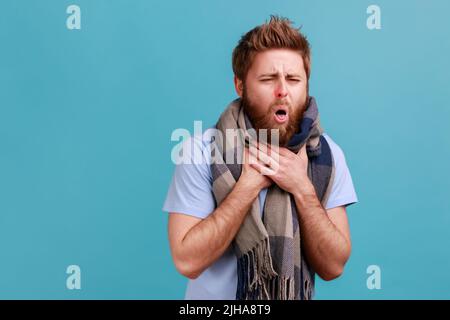 Portrait d'un homme barbu qui tousse un foulard chaud, qui se sent mal à la gorge, fièvre à haute température, symptômes de la grippe saisonnière. Studio d'intérieur isolé sur fond bleu. Banque D'Images