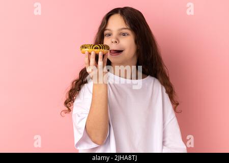 Portrait de ravissante petite fille portant un T-shirt blanc léchant délicieux beignet, cherchant avec le désir de manger un dessert sucré, montrant la langue dehors. Studio d'intérieur isolé sur fond rose. Banque D'Images