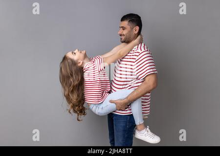 Portrait d'un père et d'une fille heureux et satisfaits en T-shirts à rayures passant du temps ensemble, en appréciant le week-end, papa tenant un enfant. Prise de vue en studio isolée sur fond gris. Banque D'Images