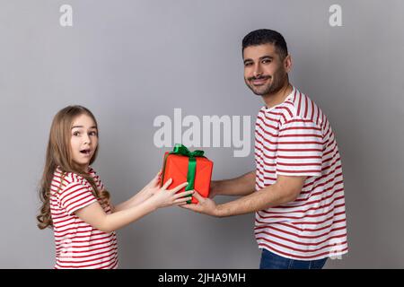 Portrait d'un père et d'une fille heureux et positif à rayures, célébrant l'anniversaire du petit enfant, papa donnant boîte cadeau à l'enfant émerveillé. T-shirts Studio d'intérieur, photo isolée sur fond gris. Banque D'Images