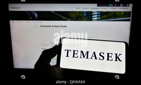 Personne tenant un téléphone portable avec le logo de la société Temasek Holdings Private Limited à l'écran en face de la page Web d'affaires. Mise au point sur l'affichage du téléphone. Banque D'Images