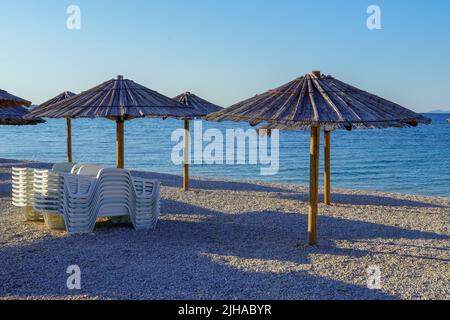 Chaises longues empilées, la mer en arrière-plan, un parasol en paille au-dessus, une image qui illustre des vacances tropicales Banque D'Images