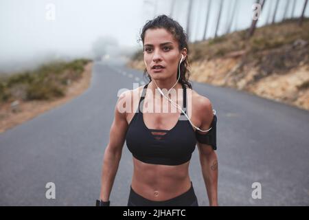 Femme musclée écoutant de la musique pendant sa séance d'entraînement du matin. Jeune femme sportive qui va courir le long d'une route brumeuse. Banque D'Images